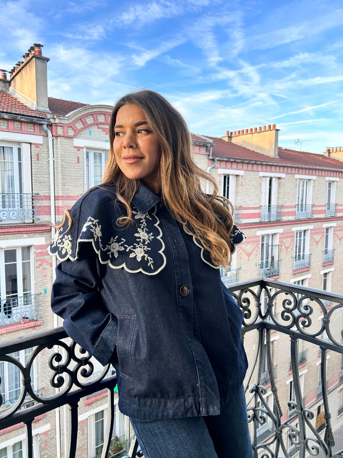 Veste en denim bleu foncé à broderies écrues portée sur un balcon parisien